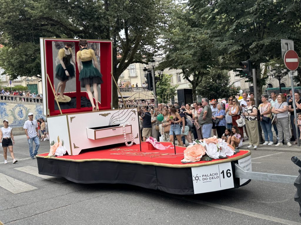 Parade float with two dancers in green tutus inside a glass-front display, atop a white and red platform, as crowds watch from the street.