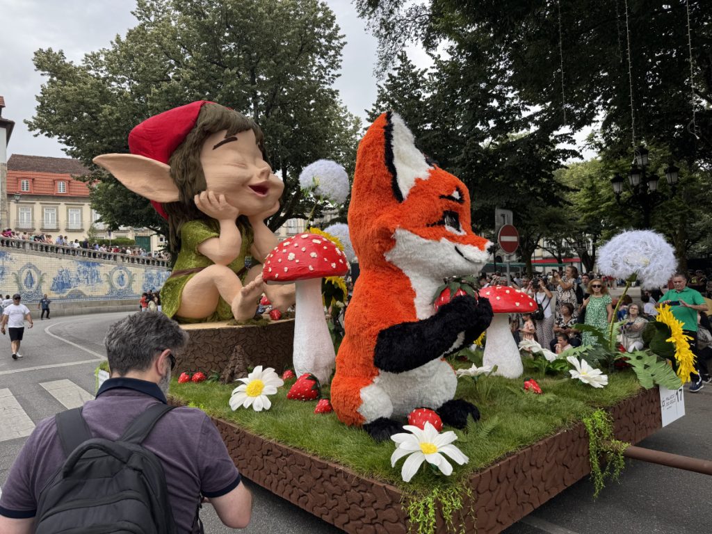 Whimsical parade float with a large elf figure and an orange fox cartoon, red mushrooms, and oversized flowers on a grassy platform, crowd watching in the background.