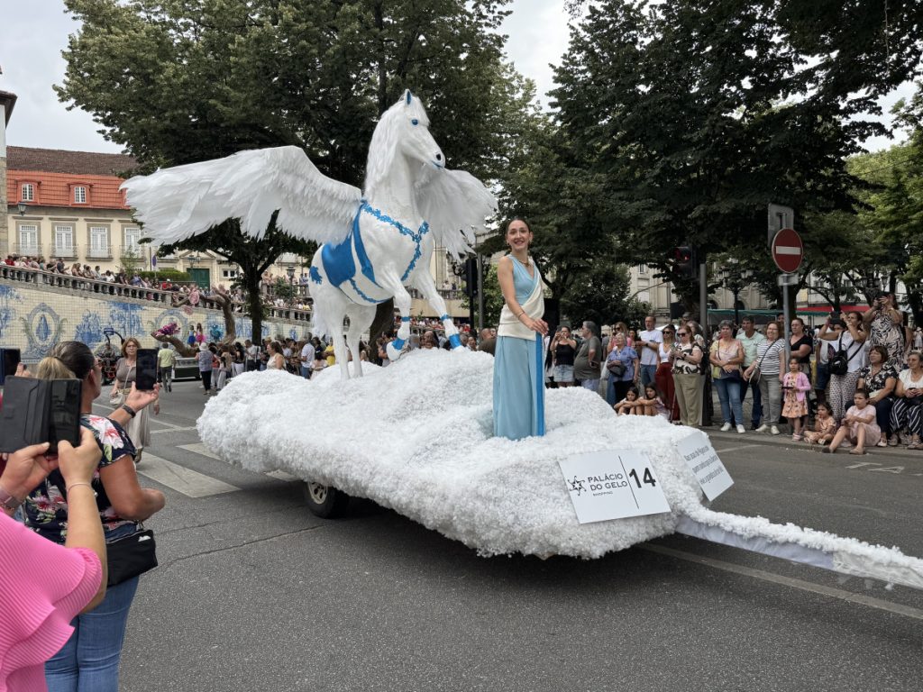 White winged horse statue on a snow-covered parade float, a woman in a blue and white gown standing beside it as crowds watch from the street edge.