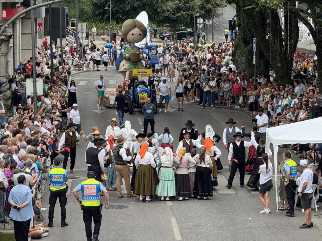Crowd watches a street parade with people in traditional regional costumes dancing in the center of the road while spectators line the sidewalks.