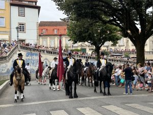 Group of riders on horses in formal attire (top hats, yellow vests) marching down a street while a crowd watches from both sides and a red flag leads the parade.