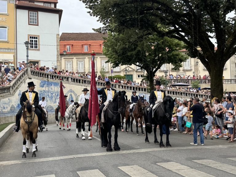 Group of riders on horses in formal attire (top hats, yellow vests) marching down a street while a crowd watches from both sides and a red flag leads the parade.