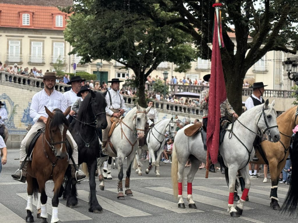 Group of riders in white shirts and hats parading on horses down a city street while crowds watch from the sides.