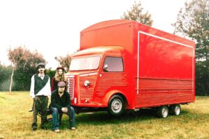 Three men in vintage clothing pose beside a bright red box truck on a grassy field.