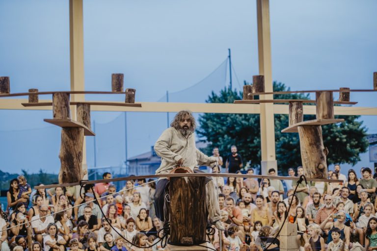 Bearded man in a beige robe stands on a tall wooden post structure, performing before a large outdoor audience.