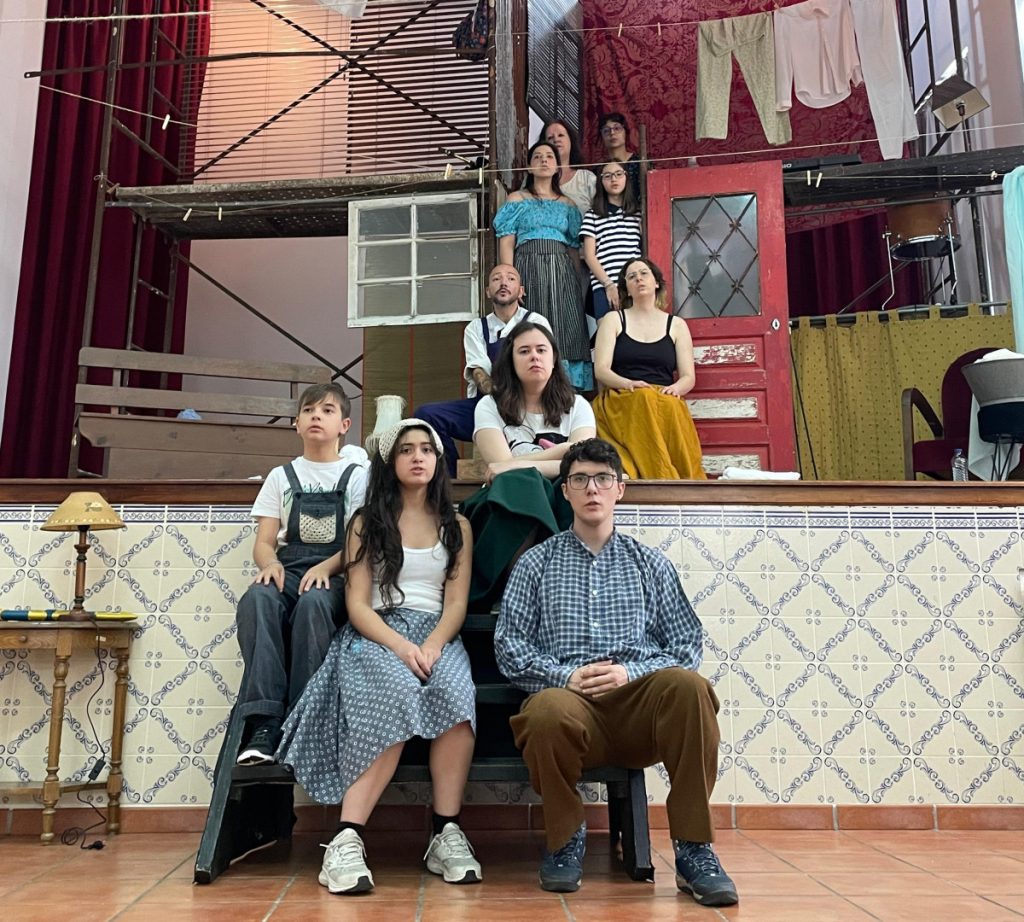 Group of young actors posing on a multi-level rehearsal set with scaffolding, a red door, and hanging laundry in the background.