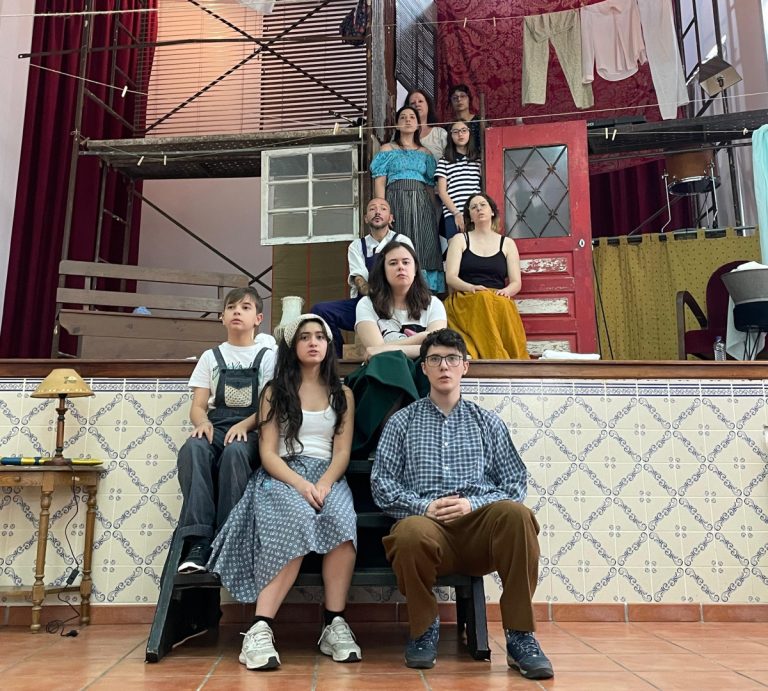 Group of young actors posing on a multi-level rehearsal set with scaffolding, a red door, and hanging laundry in the background.