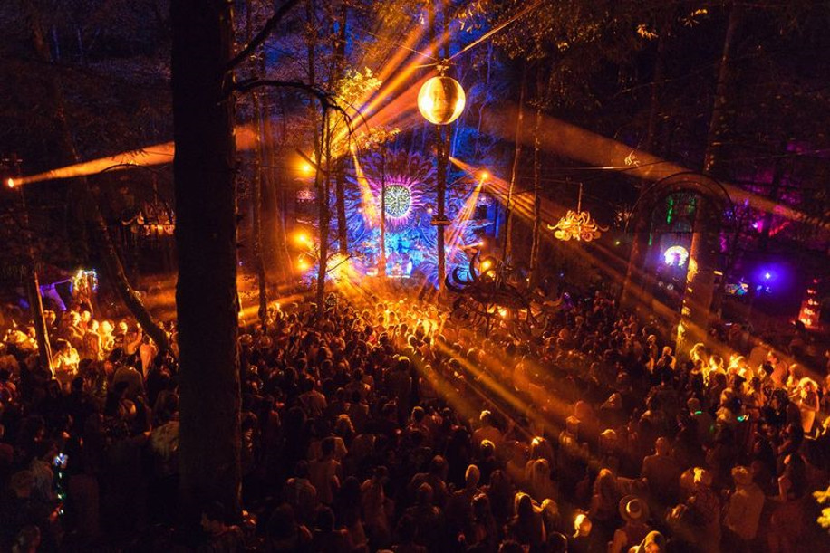 Crowded outdoor night-time dance party in a forest with colorful stage lights and a hanging disco ball menacing the crowd.