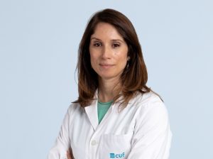 Female healthcare professional in a white lab coat, arms crossed, calm expression, light blue background. CUF logo visible on coat pocket.