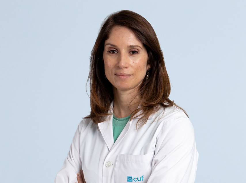 Female healthcare professional in a white lab coat, arms crossed, calm expression, light blue background. CUF logo visible on coat pocket. 10 anos Hospital Cuf Viseu: “Hoje, mais do que tratar a doença, é necessário acompanhar percursos de saúde cada vez mais complexos”