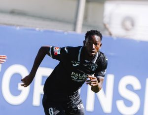 Soccer player in a black jersey sprinting on the field during a match, number 6 visible on shorts, blue advertising boards in the background