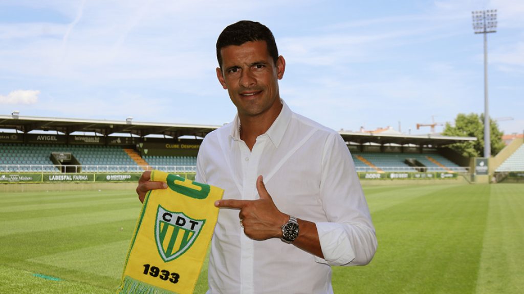 Man in white shirt on a soccer field holding a yellow CDT banner with 1933 text in a stadium. Ricardo Costa é o novo treinador do Tondela