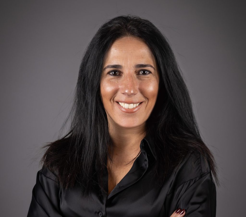 Smiling woman with long dark hair wearing a black blouse against a gray studio backdrop. Antecipar para manter as portas abertas