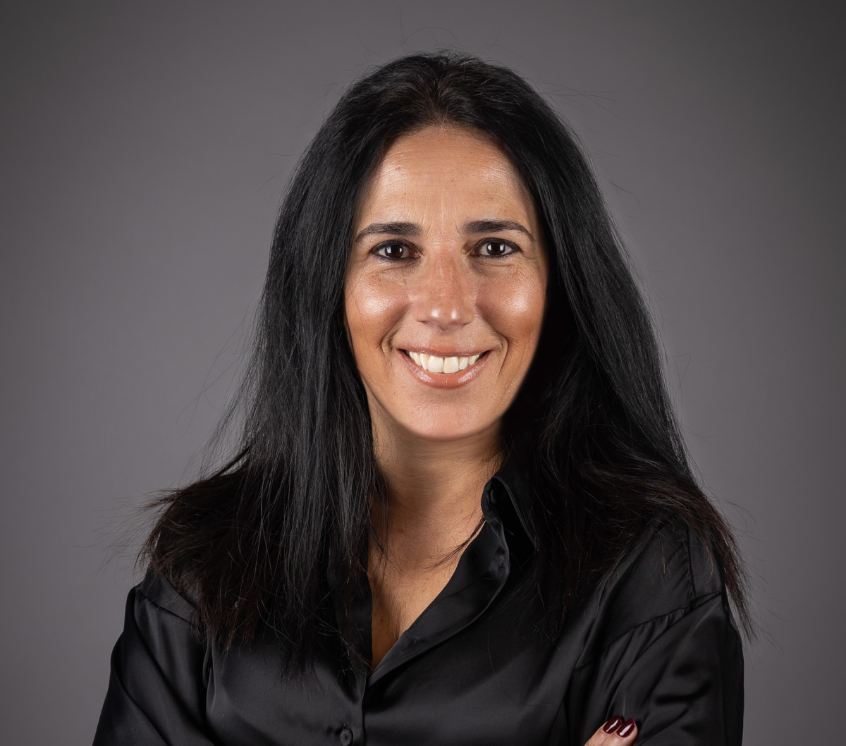 Smiling woman with long dark hair wearing a black blouse against a gray studio backdrop. Antecipar para manter as portas abertas