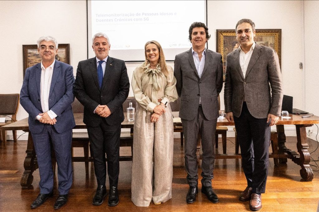 Five professionals in suits and business attire pose for a group photo in a conference room, with a projection screen behind them. Região Viseu Dão-Lafões vai alargar telemonitorização 5G em pessoas vulneráveis