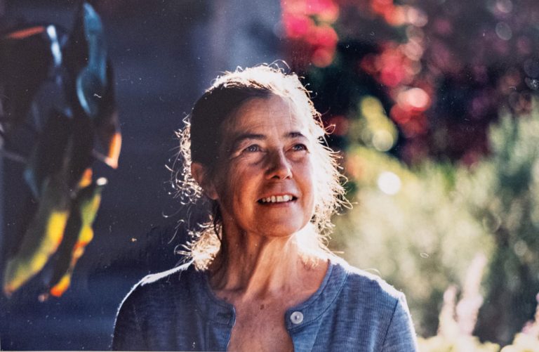 Older woman with gray hair smiling outdoors in sunlight, resembling a candid garden portrait.