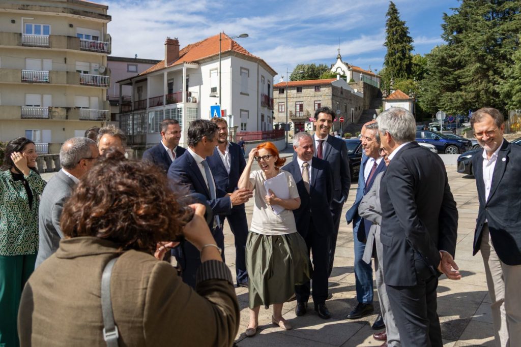 Group of professionals in suits and smart casual clothing standing on a sunny street, central woman with red hair gesturing while holding papers.