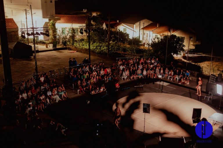 Audience seated in a semicircle around an outdoor circular stage at night, performers visible under stage lights.