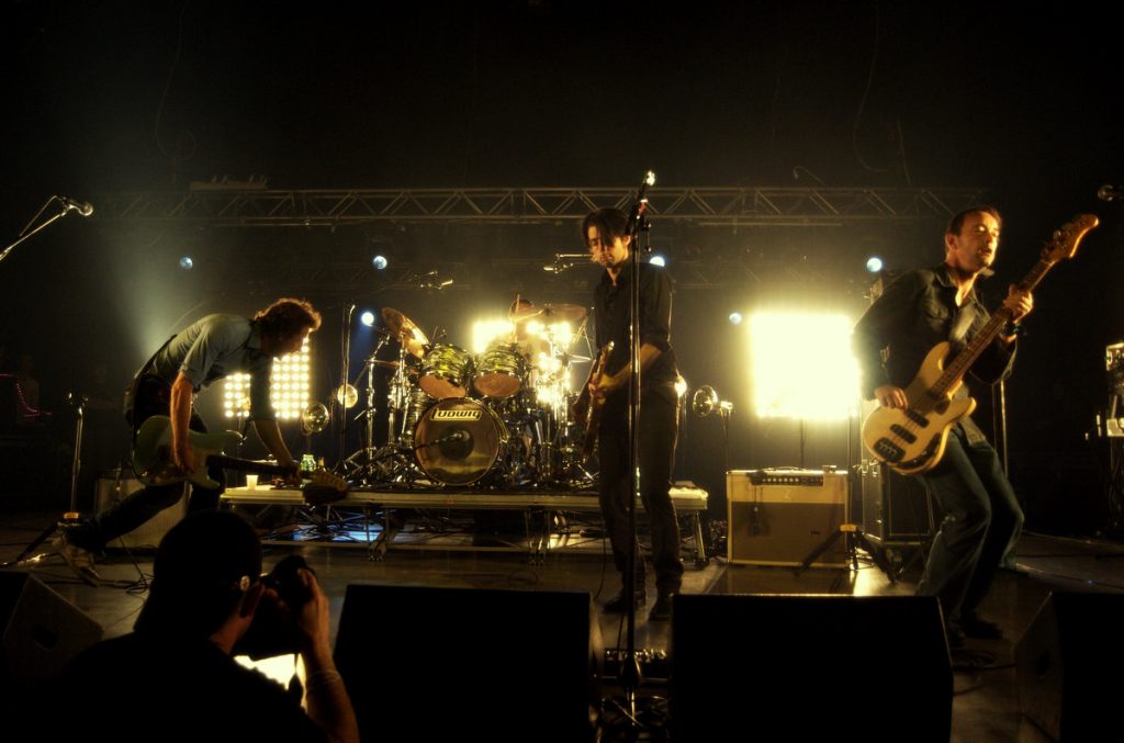 Four-piece rock band on a dark stage: left guitarist bending over, center guitarist and vocalist, drummer at back, and right bassist playing under bright backlights.
