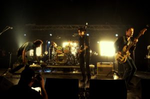 Four-piece rock band on a dark stage: left guitarist bending over, center guitarist and vocalist, drummer at back, and right bassist playing under bright backlights.
