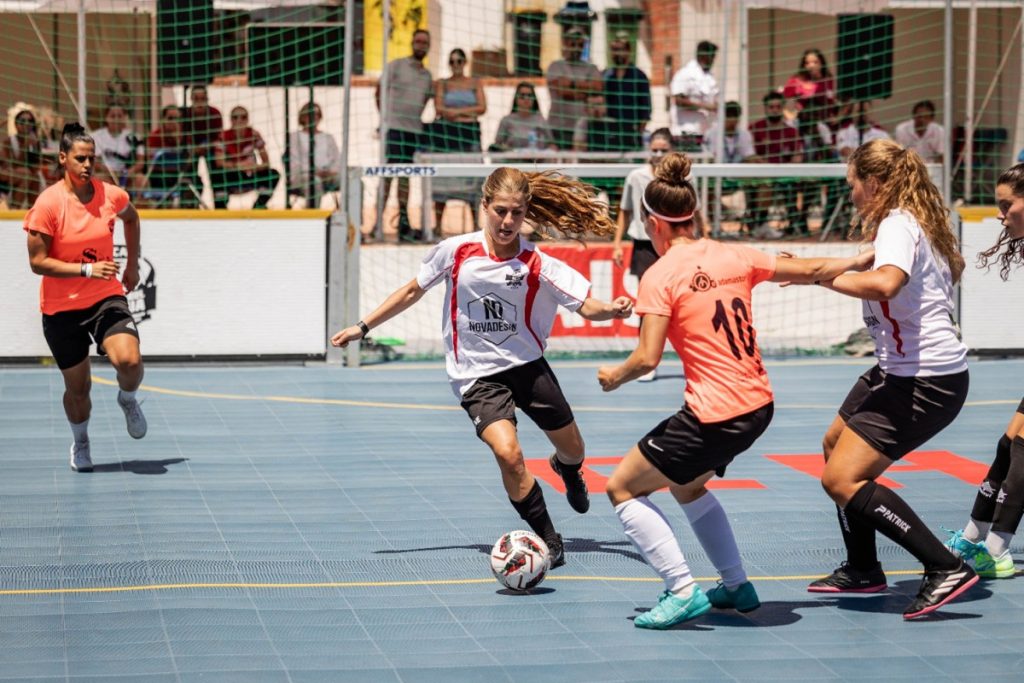 Female futsal players chase a ball on a blue outdoor court, white and orange jerseys, spectators watching behind a net.