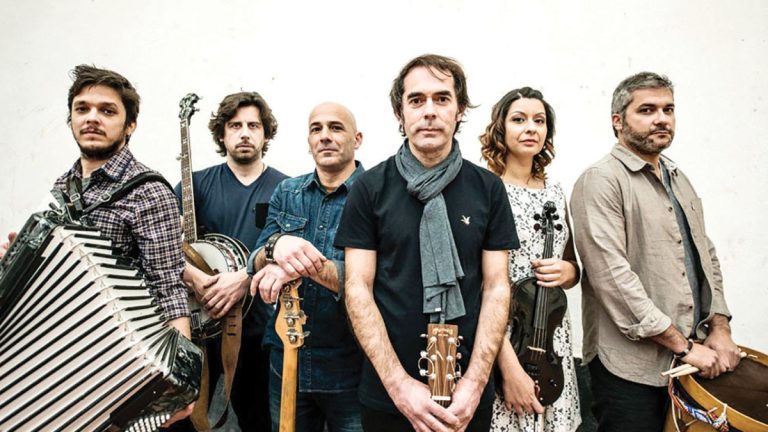 Six musicians pose with their instruments: accordion, banjo, guitars, violin, and percussion on a white backdrop.