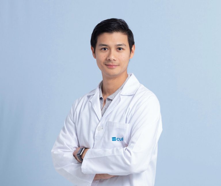 Portrait of a medical professional in a white lab coat with the CUF logo, arms crossed, smiling against a light blue backdrop.