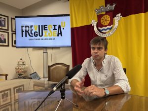 Man in a striped shirt at a press conference, speaking into a microphone on a glass table with a yellow‑red flag and crest in the background.