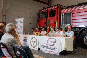 Panel discussion at a charity/event inside a fire station, with a red fire truck behind and banners on the table centerpiece.