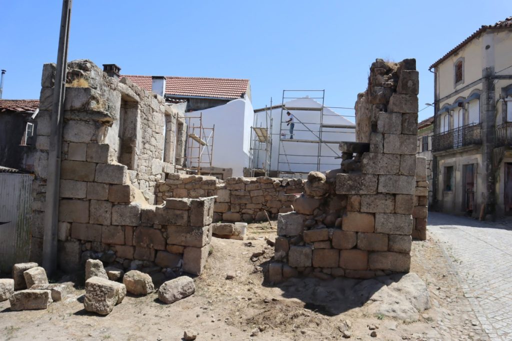 Ruinous stone walls and scattered blocks along a sunny street, with scaffolding around a white building in the background (construction site). Estudos permitem reinterpretar 500 anos de história da antiga Casa da Câmara de Aguieira, em Nelas