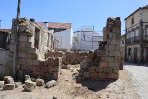 Ruinous stone walls and scattered blocks along a sunny street, with scaffolding around a white building in the background (construction site).