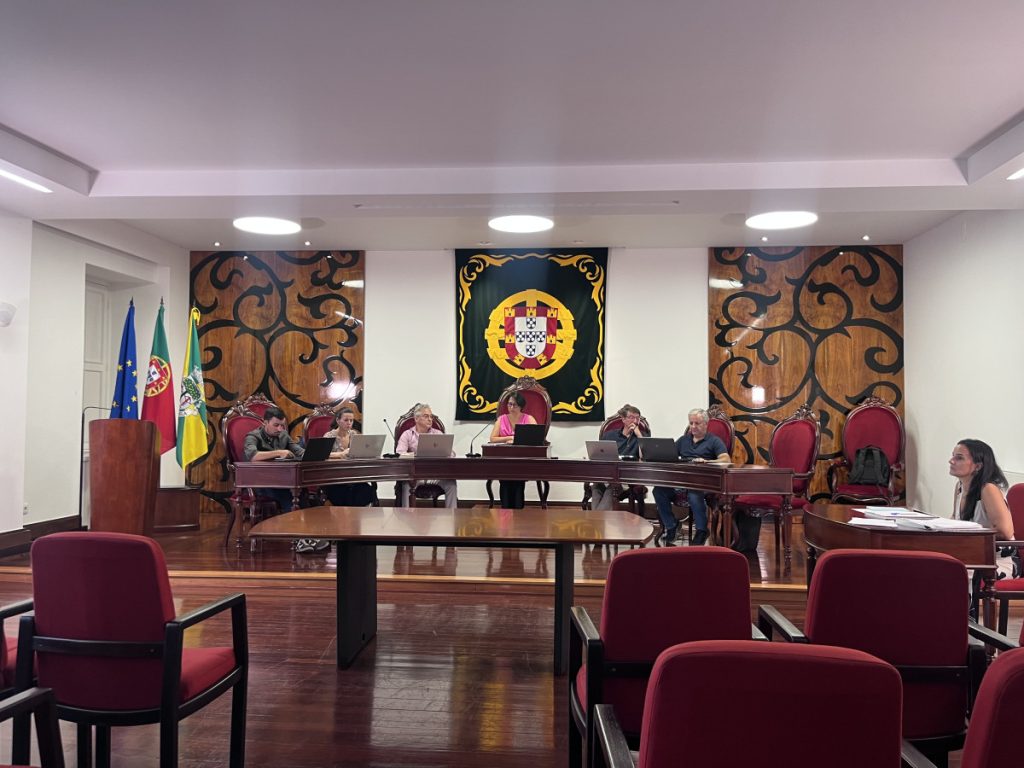 Panel of people seated at a long dark wood table in a formal meeting room with multiple laptops and flags nearby; a crest hangs on the wall behind the panel. Autarquia de Tondela prepara plano para mitigar efeitos do fogo na serra do Caramulo