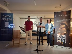 Three speakers stand on a small stage between blue and dark banners, with a microphone on a stand in front of them.