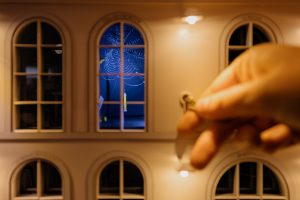 Close-up of a hand placing a tiny key into a lock on a dollhouse facade with arched windows; blue-lit interior visible through the center window with a spiderweb