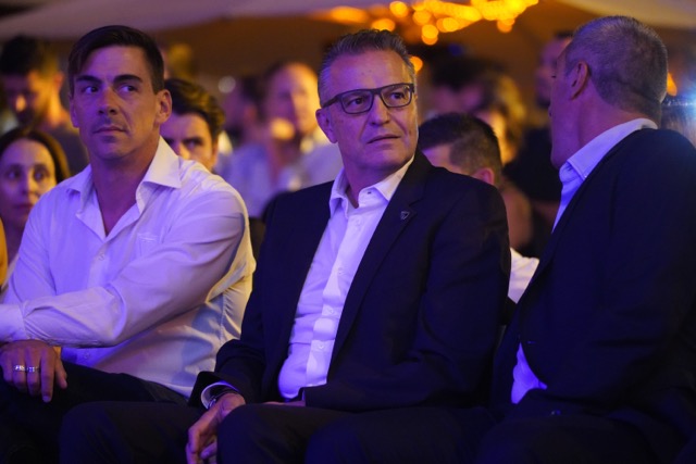 Three men in formal attire sit in the front row at an event: left in a white shirt, center in a dark suit with glasses, right facing away, with a blurred audience in warm lighting behind them. Académico de Viseu com plantel “coeso e humilde” no regresso à I Liga, 37 anos depois