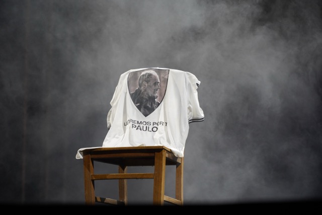 White T-shirt with a portrait printed on it, draped over a wooden chair on a foggy stage creating a memorial/tribute display.