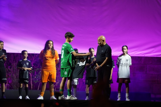 Youth soccer awards on stage: a player in green hands a jersey to a teammate while others watch under purple lights. Académico de Viseu com plantel “coeso e humilde” no regresso à I Liga, 37 anos depois