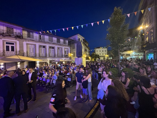 Night festival in a town square with a large crowd, string bunting, and a statue in the center. Académico de Viseu com plantel “coeso e humilde” no regresso à I Liga, 37 anos depois