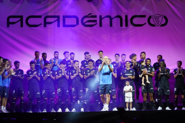 Team of soccer players on a stage, clapping as a man in a blue shirt speaks into a microphone; a child stands beside him against a purple-lit backdrop.