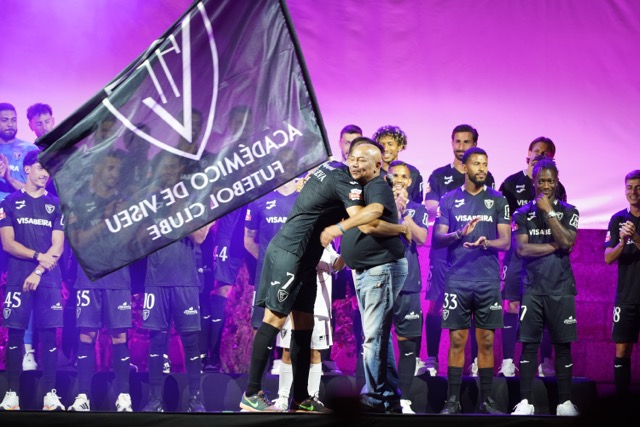Soccer team in purple uniforms on a stage celebrating; two men hug in the center while a large club flag waves in the foreground.