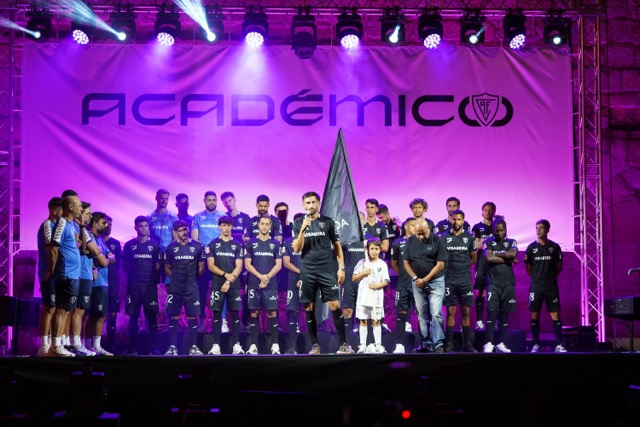 A group of soccer players on a brightly lit stage with a banner reading 'ACADEMICO' behind, a man speaks at center flanked by a child and staff.