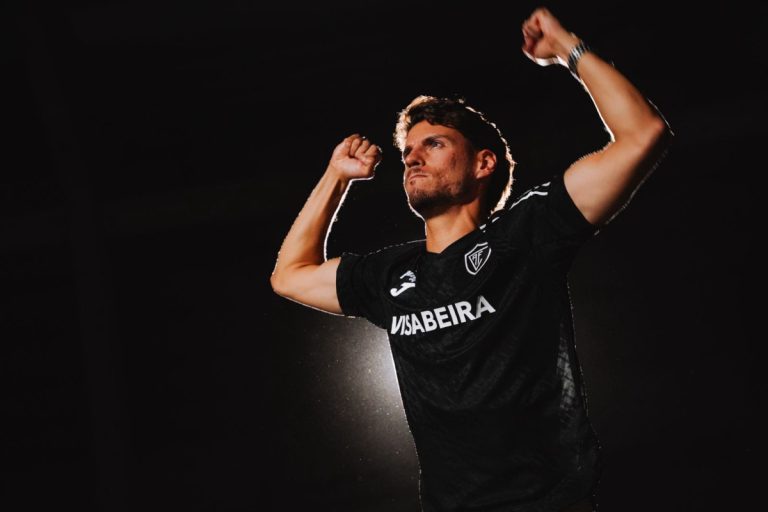Soccer player in a black jersey raises both fists in celebration against a dark background, lighting highlighting his silhouette.