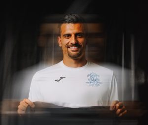 Smiling man in a white sports jersey with a crest, holding a banner in a blurred background.