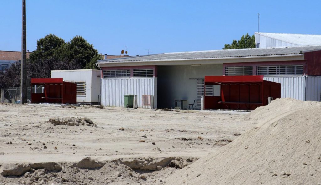 Dusty construction site with sand piles, red storage sheds, and gray metal warehouses under a clear blue sky. Nelas avança com primeiro relvado sintético municipal que deverá ficar concluído em setembro