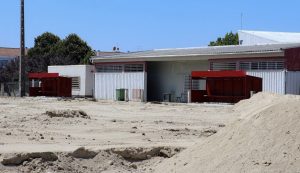 Dusty construction site with sand piles, red storage sheds, and gray metal warehouses under a clear blue sky.