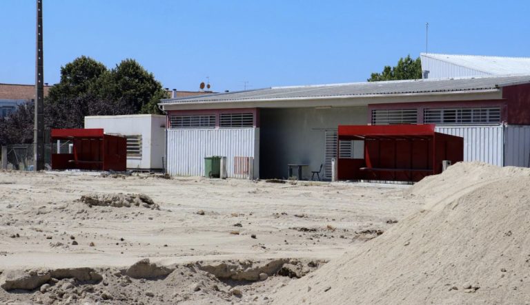 Dusty construction site with sand piles, red storage sheds, and gray metal warehouses under a clear blue sky.