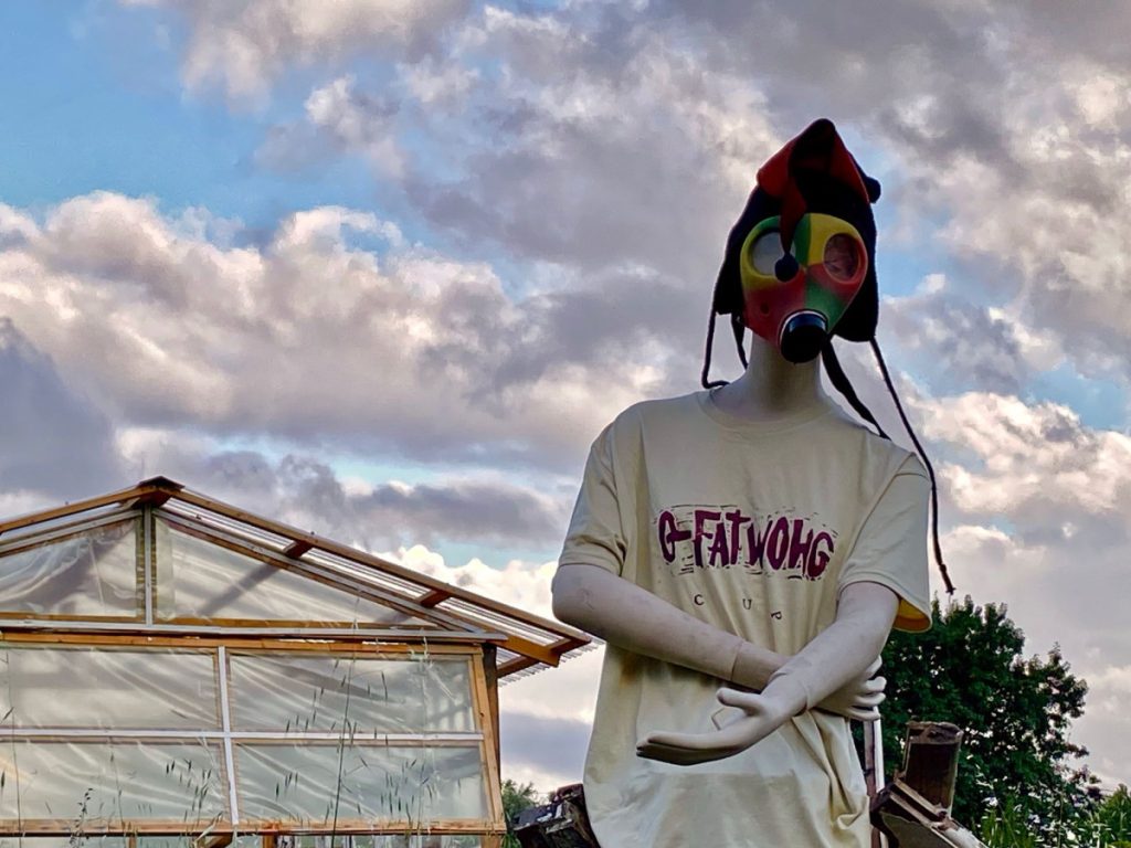 Life-sized mannequin outdoors wearing a colorful gas mask and a loose beige T‑shirt, near a glass greenhouse. Suppressed windswept clouds in the sky enhance the surreal scene.
