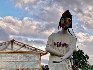 Life-sized mannequin outdoors wearing a colorful gas mask and a loose beige T‑shirt, near a glass greenhouse. Suppressed windswept clouds in the sky enhance the surreal scene.