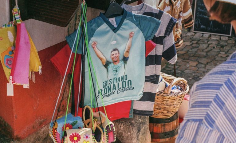 Outdoor market stall displaying clothing and colorful bags; a Ronaldo-themed blue T-shirt is central.