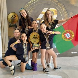 Six young dancers pose with medals and a large trophy in front of a dance competition backdrop, Portugal flag visible behind them.